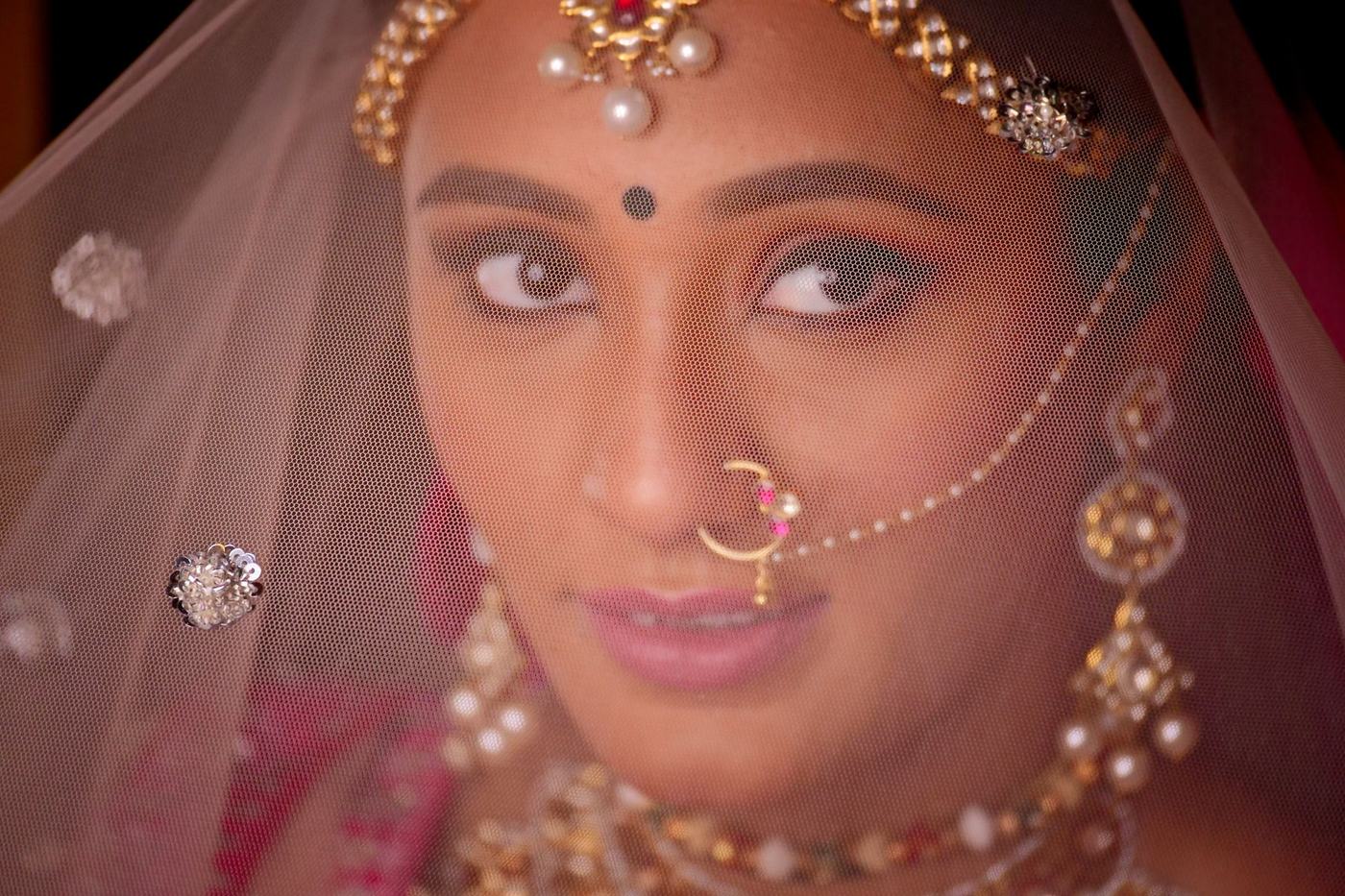 Bride's eyes seen through a sheer red veil, nose ring and maang tikka catching the light