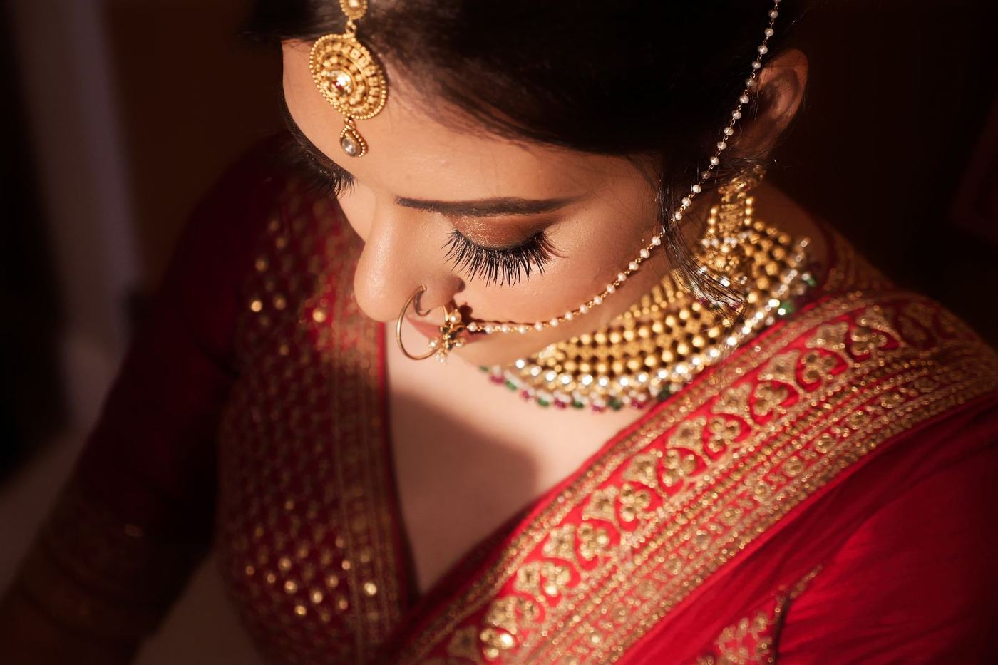 Bride in a red and gold sari looking down, nose ring chain across her cheek