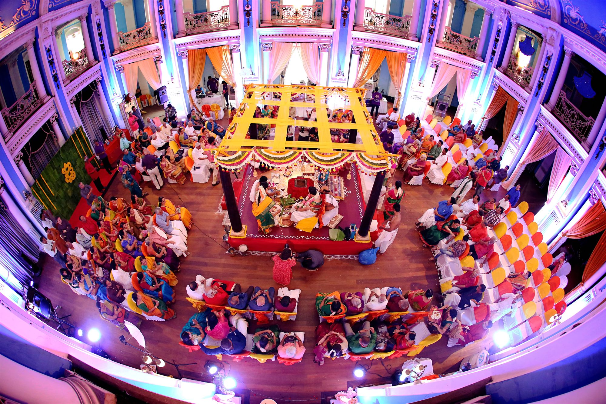 The whole ceremony hall seen at once, families circling the mandap