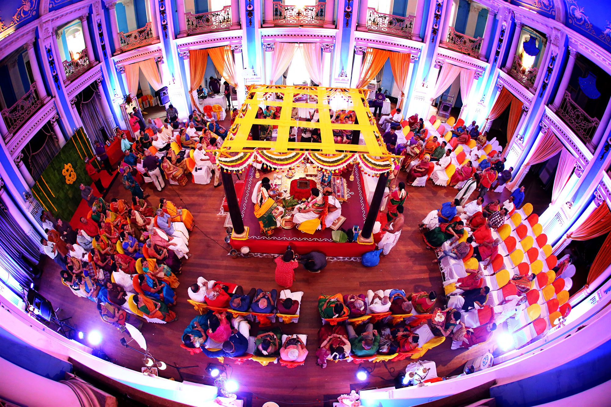 The whole ceremony hall seen at once, guests circling the mandap below