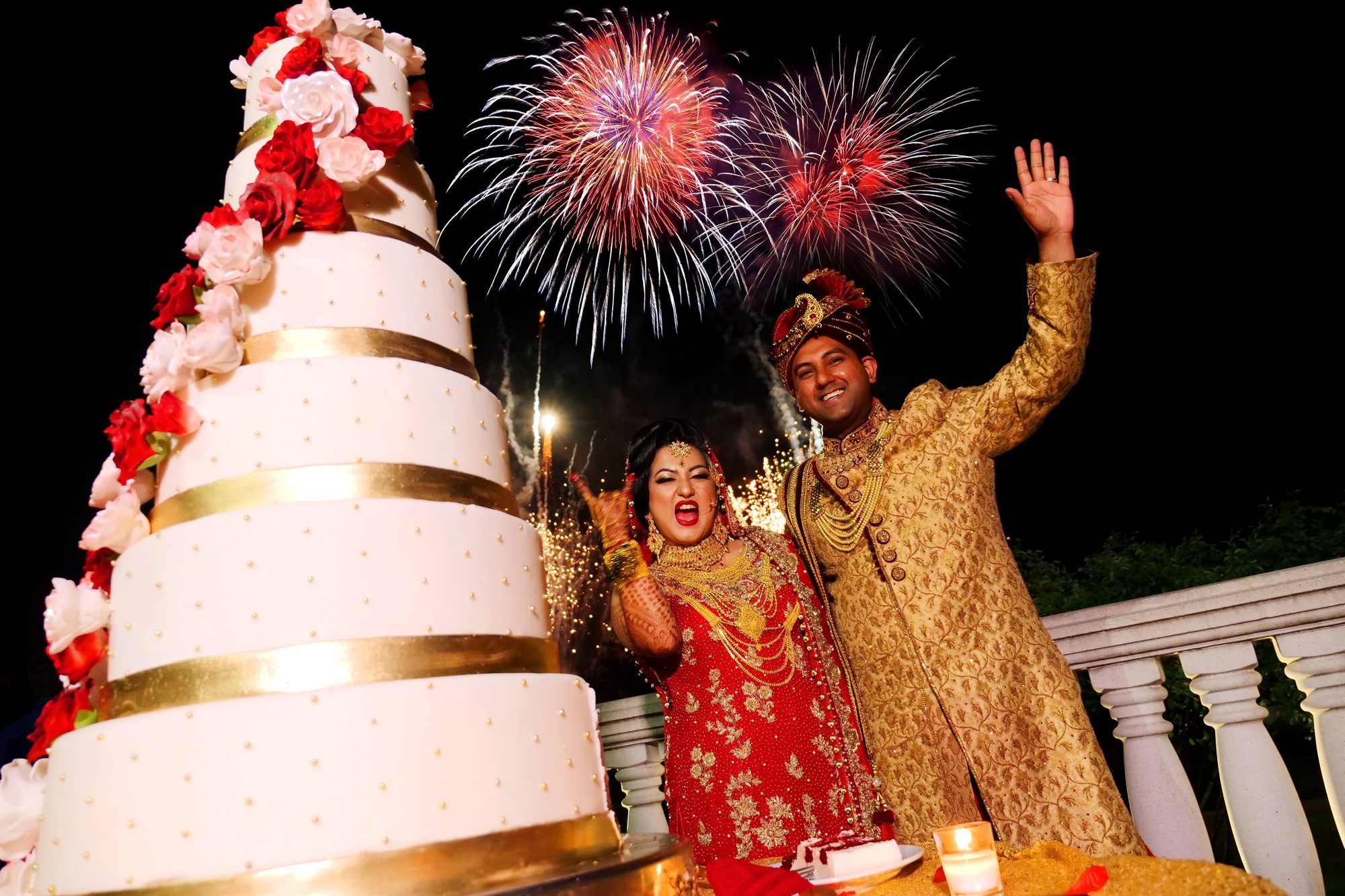 Couple raising their hands beside the cake as fireworks open behind them