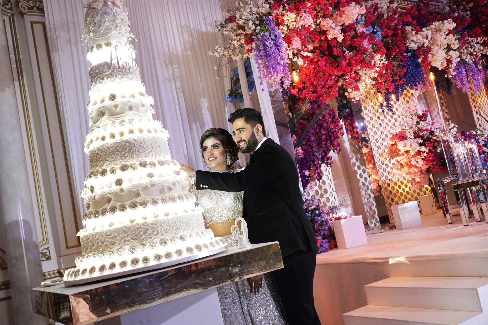 Couple cutting a tall tiered cake against a wall of flowers