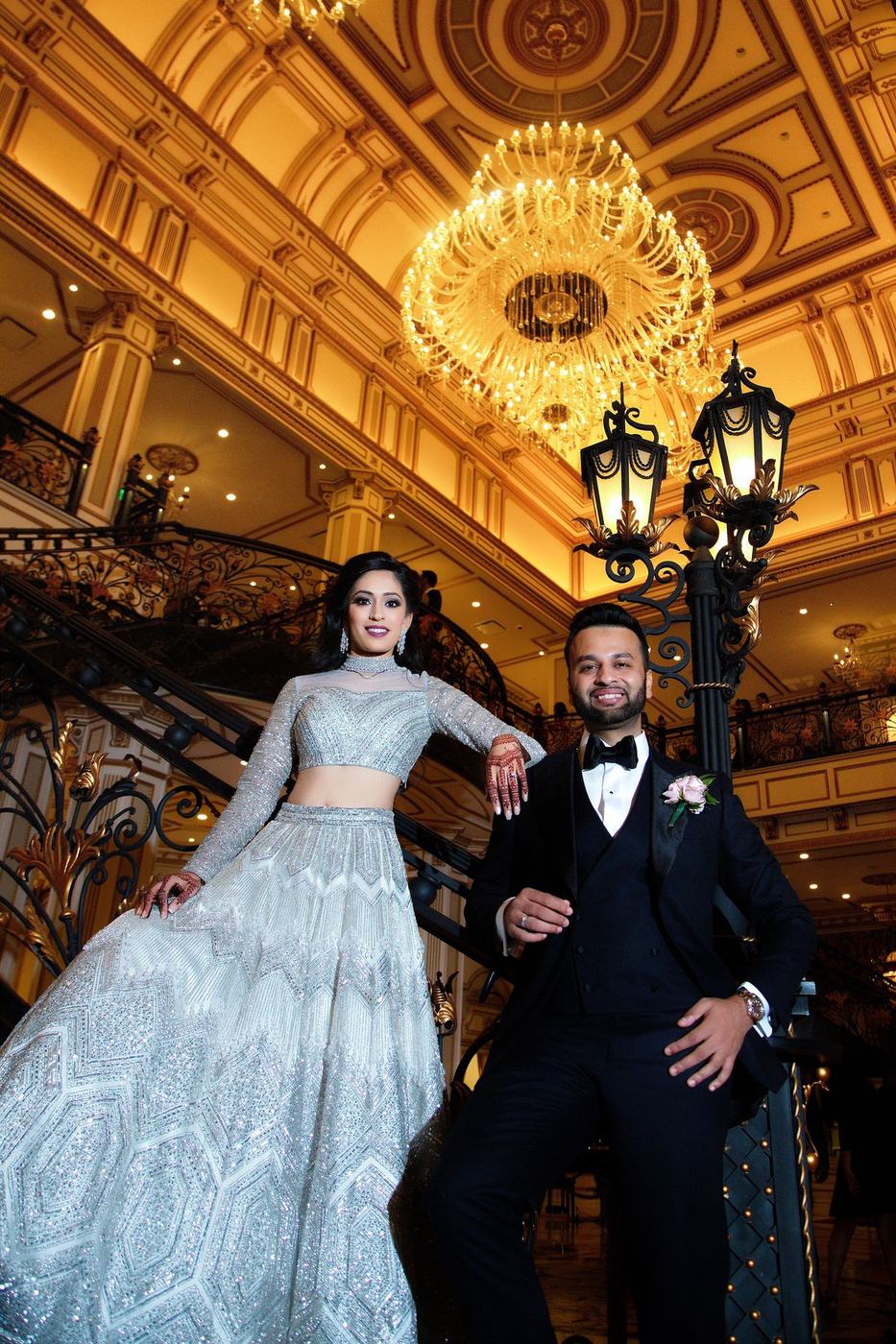 Couple in an ice-blue gown and black tuxedo at the foot of a gilded ballroom staircase