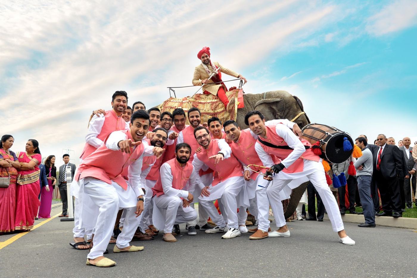 Groom on an elephant above his groomsmen in coral, under a wide sky